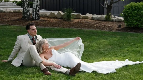 A bride and groom sitting on a grassy lawn, with the bride holding her veil.