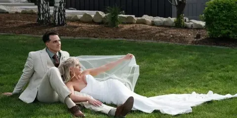 A bride and groom sitting on a grassy lawn, with the bride holding her veil.