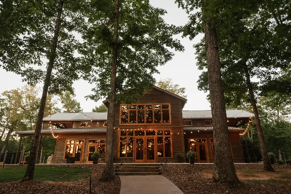 Exterior view of a large wooden event venue with string lights, surrounded by tall trees.