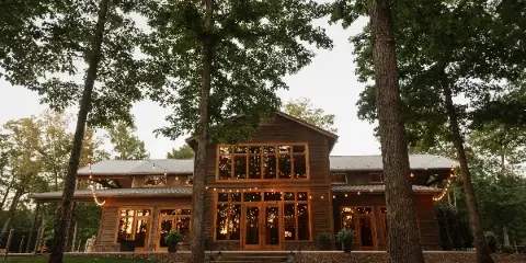 Exterior view of a large wooden event venue with string lights, surrounded by tall trees.