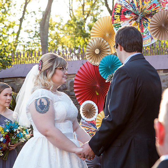 A bride and groom holding hands during an outdoor wedding ceremony with a colorful paper fan backdrop.