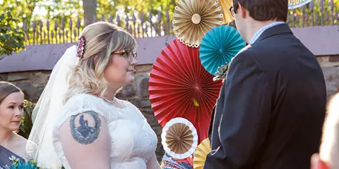 A bride and groom holding hands during an outdoor wedding ceremony with a colorful paper fan backdrop.