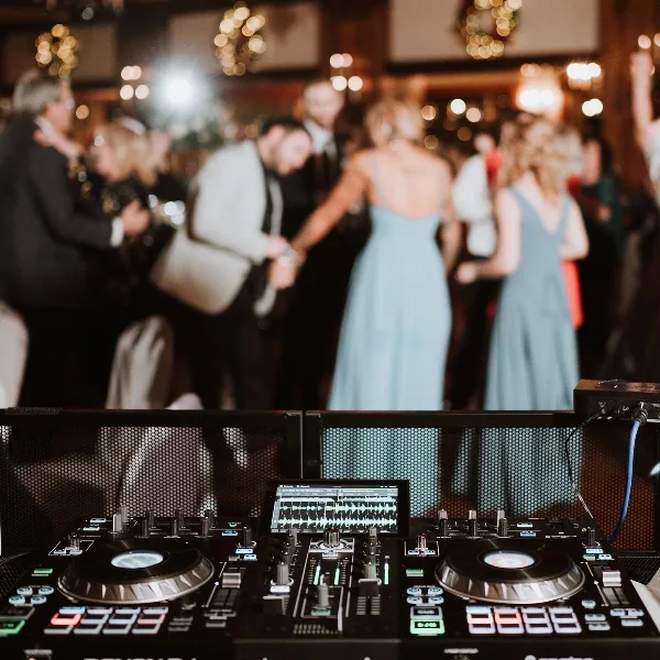 Professional DJ equipment setup in the foreground with a blurred background of wedding guests dancing at a reception.