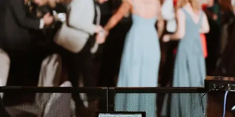 Professional DJ equipment setup in the foreground with a blurred background of wedding guests dancing at a reception.