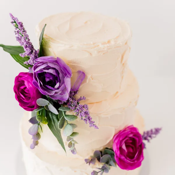 A close-up of a two-tiered white frosted cake decorated with purple and pink roses and greenery.