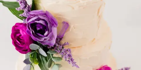 A close-up of a two-tiered white frosted cake decorated with purple and pink roses and greenery.