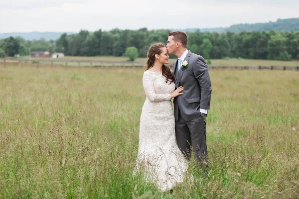 A bride and groom standing in a grassy field with a wooden fence and farm buildings in the background.