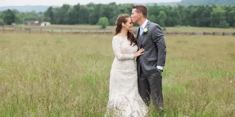 A bride and groom standing in a grassy field with a wooden fence and farm buildings in the background.