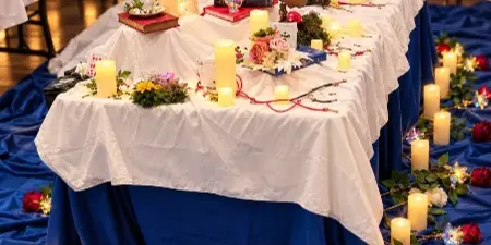 A long event table decorated with white tablecloths, blue draping, pillar candles, and floral arrangements including roses and greenery.