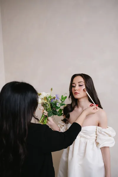 A makeup artist applying makeup to a bride in a white dress holding a floral bouquet.