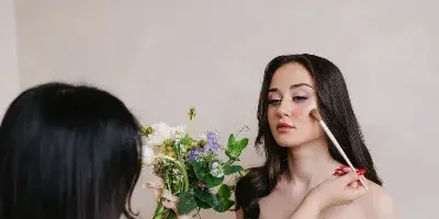 A makeup artist applying makeup to a bride in a white dress holding a floral bouquet.