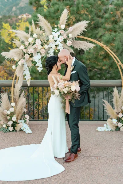 A bride and groom embracing in front of a floral wedding arch with pampas grass and roses.
