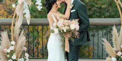 A bride and groom embracing in front of a floral wedding arch with pampas grass and roses.