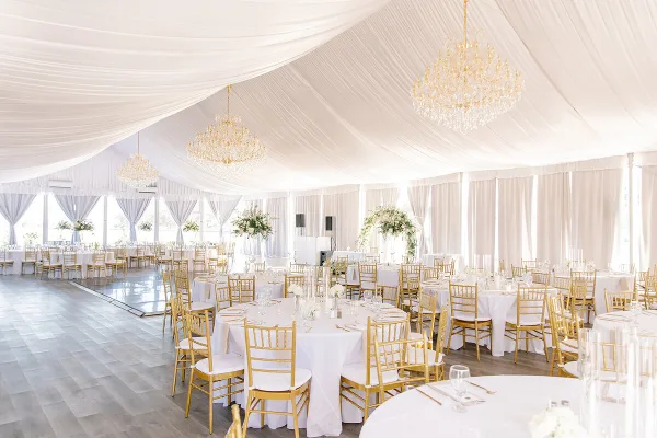 A luxury wedding reception venue featuring white draping, gold chandeliers, and round tables with gold chiavari chairs.