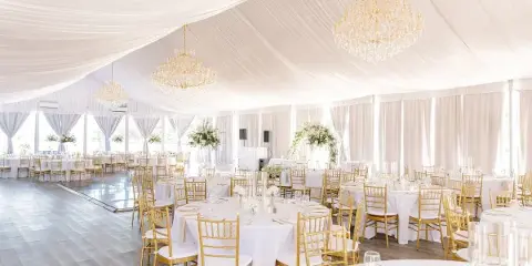 A luxury wedding reception venue featuring white draping, gold chandeliers, and round tables with gold chiavari chairs.