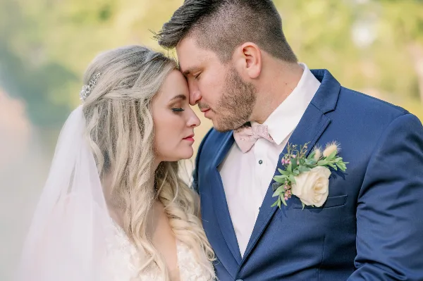 A bride and groom in formal wedding attire standing close together outdoors, with the groom wearing a blue suit and the bride in a white dress and veil.