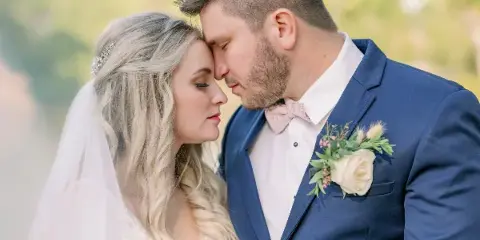 A bride and groom in formal wedding attire standing close together outdoors, with the groom wearing a blue suit and the bride in a white dress and veil.