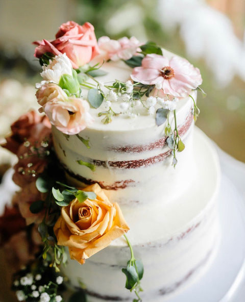 A two-tier semi-naked wedding cake decorated with fresh pink and yellow roses and greenery.