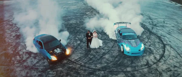 A bride and groom standing between two cars performing burnouts on a paved surface.