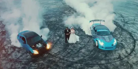 A bride and groom standing between two cars performing burnouts on a paved surface.