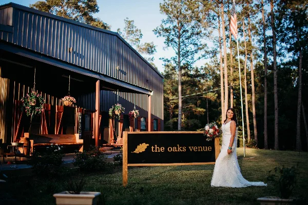 A bride in a white wedding dress stands next to a wooden sign reading 'the oaks venue' in front of a rustic metal barn building surrounded by pine trees.