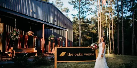 A bride in a white wedding dress stands next to a wooden sign reading 'the oaks venue' in front of a rustic metal barn building surrounded by pine trees.