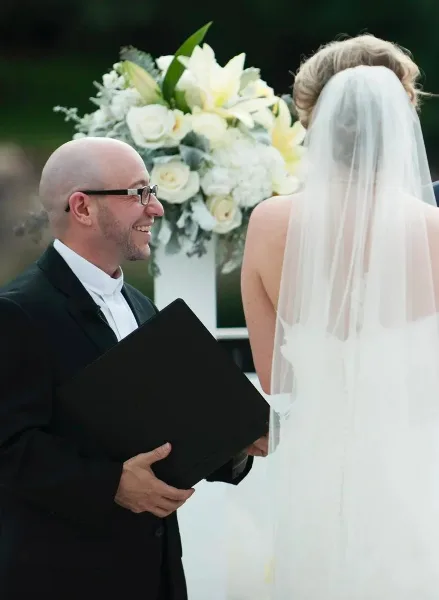 A wedding officiant in a black suit and white clerical collar smiles while facing a bride in a white wedding dress and veil during a ceremony.