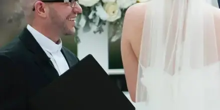A wedding officiant in a black suit and white clerical collar smiles while facing a bride in a white wedding dress and veil during a ceremony.