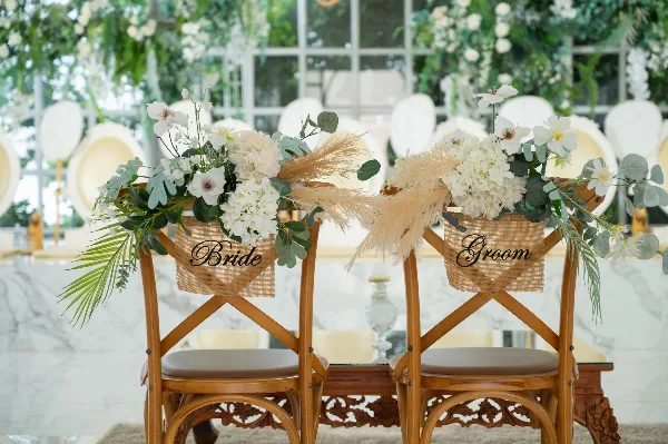 Two wooden chairs decorated with white flowers and burlap signs reading 'Bride' and 'Groom' in a wedding venue setting.