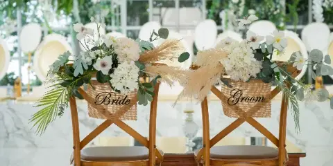 Two wooden chairs decorated with white flowers and burlap signs reading 'Bride' and 'Groom' in a wedding venue setting.