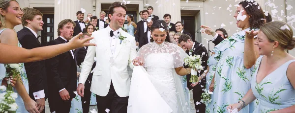 A bride and groom walking out of a building surrounded by their wedding party and falling flower petals.