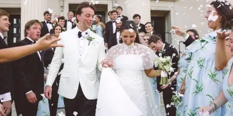 A bride and groom walking out of a building surrounded by their wedding party and falling flower petals.