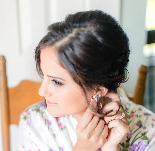 A woman with an elegant updo hairstyle and professional makeup, wearing a floral robe and putting on an earring.