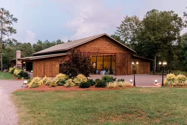 Exterior view of a wooden barn wedding venue with a covered porch, landscaped shrubs, and surrounding trees.