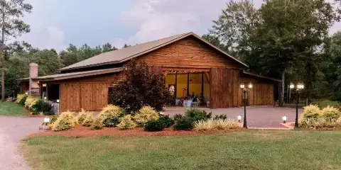 Exterior view of a wooden barn wedding venue with a covered porch, landscaped shrubs, and surrounding trees.