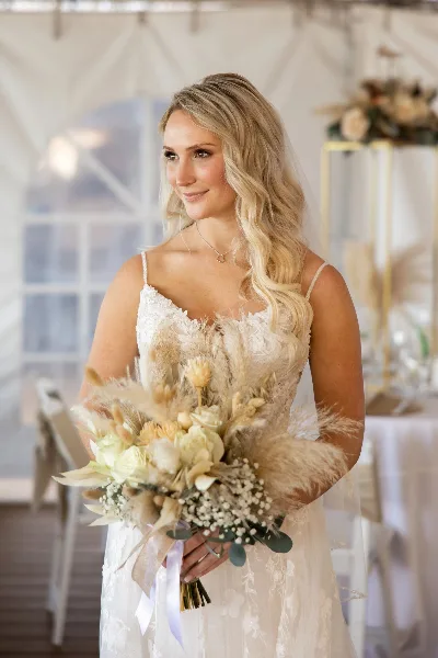 A bride in a white lace dress holding a large, textured wedding bouquet with pampas grass and white flowers.