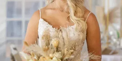 A bride in a white lace dress holding a large, textured wedding bouquet with pampas grass and white flowers.