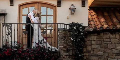 A bride and groom kissing on a balcony with floral arrangements and a stone wall.