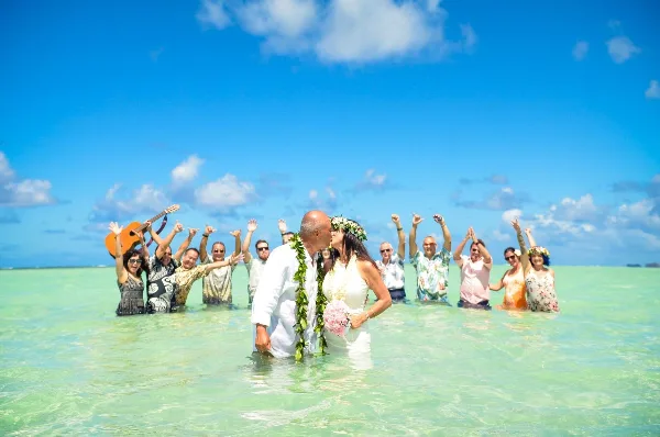 A bride and groom sharing a kiss in a shallow, turquoise ocean during a destination wedding ceremony, surrounded by guests and a musician.