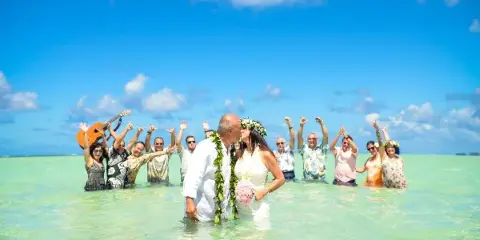 A bride and groom sharing a kiss in a shallow, turquoise ocean during a destination wedding ceremony, surrounded by guests and a musician.