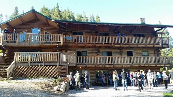 Exterior view of a two-story log cabin lodge with wooden balconies and a group of people gathered on the front patio.