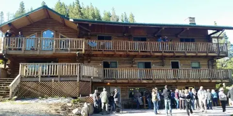 Exterior view of a two-story log cabin lodge with wooden balconies and a group of people gathered on the front patio.