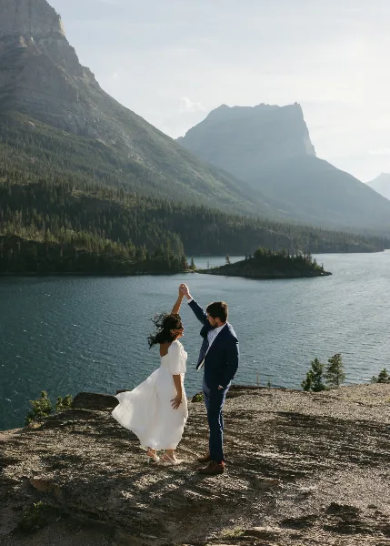 A bride and groom dancing on a rocky outcrop overlooking a mountain lake.