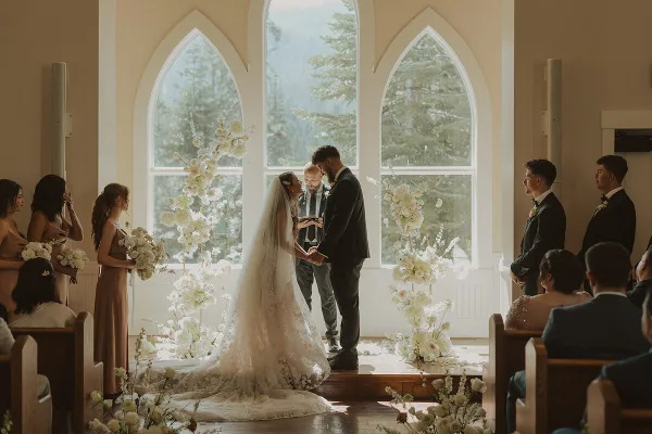 A bride and groom exchanging vows during a wedding ceremony in a chapel with large arched windows and floral arrangements.
