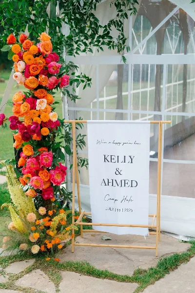 A tall, colorful floral arrangement with orange and pink roses and greenery next to a wedding banner reading 'Kelly & Ahmed'.