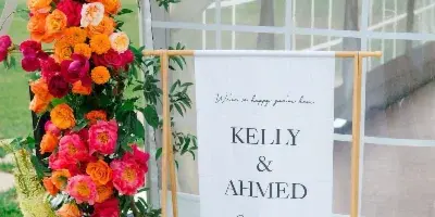A tall, colorful floral arrangement with orange and pink roses and greenery next to a wedding banner reading 'Kelly & Ahmed'.