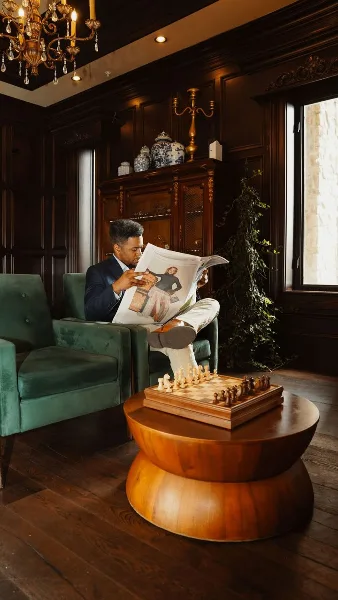 A man in a suit reads a newspaper in a wood-paneled room with a chessboard on a table and a green velvet chair.