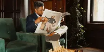 A man in a suit reads a newspaper in a wood-paneled room with a chessboard on a table and a green velvet chair.