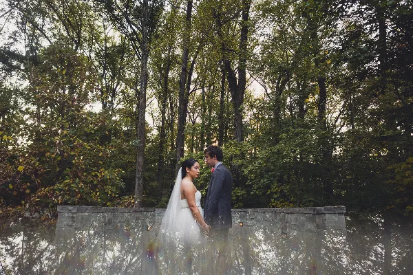 A bride and groom standing together in a wooded area near a stone wall, captured in a professional wedding photograph.