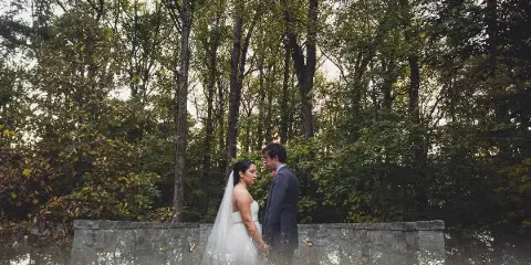 A bride and groom standing together in a wooded area near a stone wall, captured in a professional wedding photograph.
